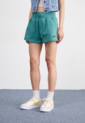 Turkise Nike sweatshorts med snor i taljen, prydet med et sort logo. Sættes sammen med cremefarvede sneakers og hvide sokker på et blåt tæppe.