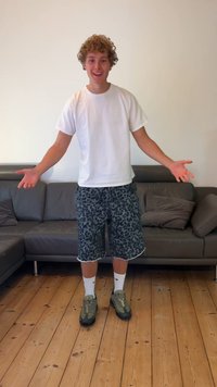 Person, die ein weißes T-Shirt, graue Leopardenmuster-Shorts, weiße Socken und grüne Schuhe trägt, steht in einem Wohnzimmer mit einer grauen Sofas.