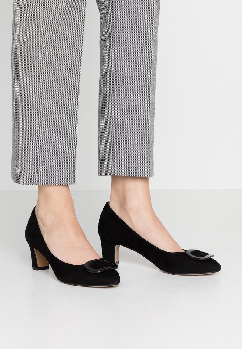 Chaussures en daim noir à talon moyen avec boucle décorative portées avec un pantalon court gris à motifs, présentées sur un sol blanc lisse.