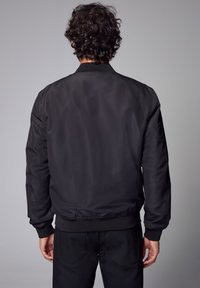 Giacca bomber nera con una texture liscia, colletto e vita a costine. Presenta polsini elasticizzati e un design semplice e lineare.