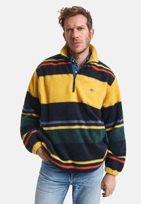 Mann in einem mehrfarbigen gestreiften Fleece-Pullover mit Halb-Reißverschluss und hellblauen Jeans, steht mit einer Hand in der Tasche vor einem neutralen Hintergrund.
