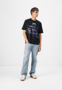 Sort T-shirt med farverig racerbil grafik og teksten "BWT ALPINE". Matchet med lyseblå jeans og hvide sneakers med brune såler.