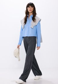Camicia azzurro chiaro con bottoni, maglione grigio appoggiato sulle spalle, pantaloni grigi a righe sottili e sneakers bianche. Cappello crema tenuto in mano.