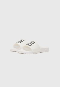 Witte slipper sandalen met een rubberen bovenkant met zwarte tekst. De zool heeft een textuur en is licht geribbeld voor grip. Eenvoudig en open ontwerp.