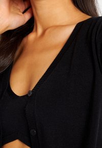 Svart stickad cardigan med djup V-ringning, featuring tre knappar framtill och ribbade kanter. Mjuk textur, delvis lager-på-lager över en svart topp.