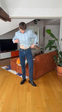 Homme en chemise rayée et jeans levant sa chemise pour montrer son ventre, debout dans un salon moderne avec un canapé orange et une grande plante.