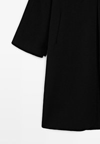 Sort langærmet frakke med lige snit, vist mod en hvid baggrund.