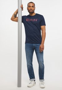 Navy blue cotton t-shirt with "MUSTANG" logo in red, paired with light blue denim jeans and white sneakers, standing against a pole.