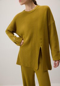 Kollane soonikoz särgi kudum, millel on lõdva lõikega, külgsplitid ja nööbid varrukatel. Ühendatud sobivate soonikute kudumipükstega.