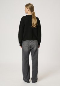 Schwarzer, strukturierten Pullover mit rundem Ausschnitt, kombiniert mit hoch taillierten, locker sitzenden grauen Jeans. Glattes Haar ist zu einem Pferdeschwanz frisiert.