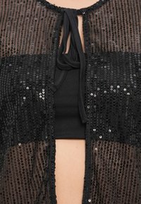 Capa transparente preta com lantejoulas, com fecho de laço, apresentando um top preto liso por baixo. Textura transparente e lantejoulas brilhantes visíveis.