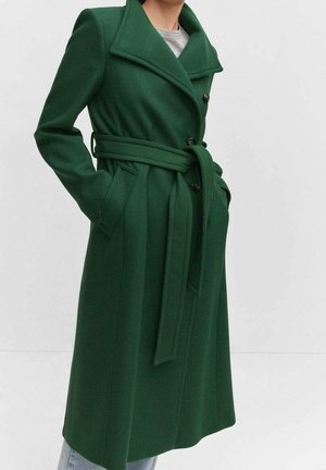 Femme portant un long manteau vert foncé ceinturé, avec de larges revers, boutonné devant et les mains dans les poches latérales, sur un fond clair uni.