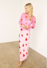 Γυναίκα που φοράει ροζ διάφανο μακρυμάνικο τοπ και ταιριαστή φούστα με κόκκινα και χρυσά μοτίβα καρδιών, στέκεται σε τσιμεντένιο πάτωμα.