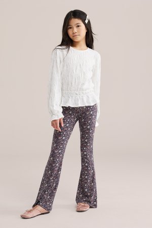 Meitene ar gariem tumšiem matiem un pērļu matu sprādzi, tērpusies baltā teksturētā garroku kreklā un ziedu raksta zvīņotās biksēs ar rozā kurpēm.