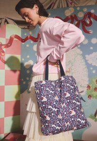 Rózsaszín pulóvert és krémszínű szoknyát viselő nő tengerészkék virág- és állatmintás vászontáskát tart színes, játékos háttér előtt.