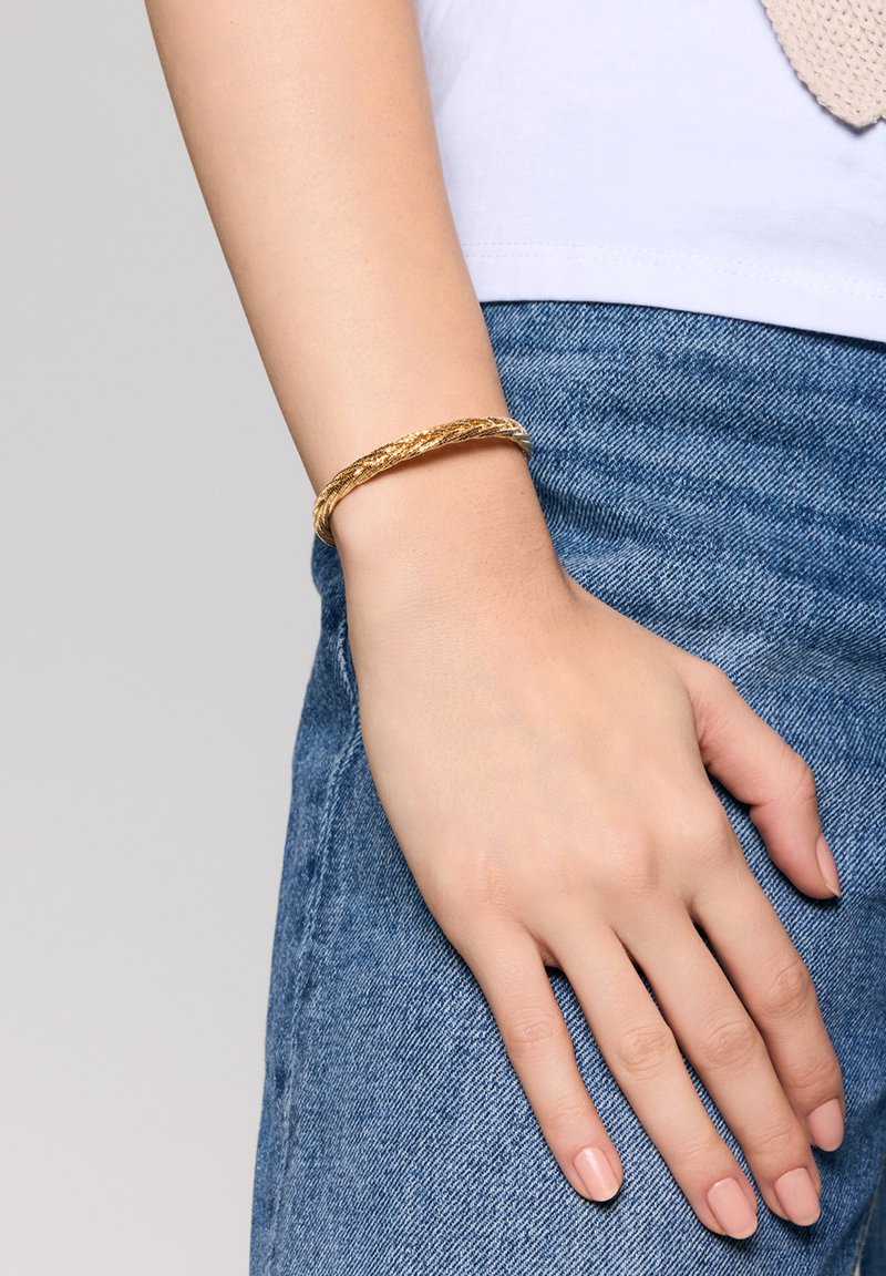 Złota pleciona bransoletka na nadgarstku. Gładka tekstura o zakrzywionym, okrągłym kształcie. Noszona na jasnej bluzce i niebieskich dżinsach.