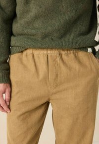 Une personne portant un pantalon en velours côtelé beige et un pull en tricot vert avec une étiquette de marque visible sur la couture latérale du pull.