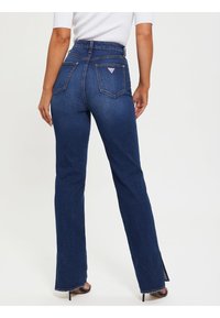 Jeans de talle alto de color azul oscuro con piernas acampanadas, que cuentan con bolsillos traseros y un pequeño logo en la cinturilla. Textura de denim suave.