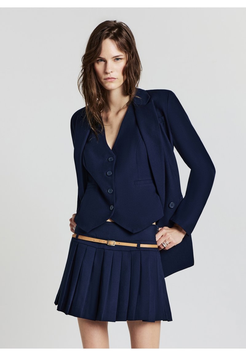 Ensemble de costume marine comprenant un blazer ajusté et une mini-jupe plissée, avec un haut en v et une ceinture en tan. Tissu lisse et structuré.