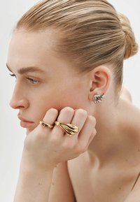 Joyas de oro y plata con diseños texturizados. Un anillo de oro con una piedra roja y un anillo escultórico de oro. Se puede ver un pendiente de plata en la oreja.