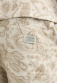 Un tissu beige à motifs présente des dessins complexes avec des feuilles et des motifs floraux. Une étiquette cousue identifie la marque "Petrol Ind & Co."