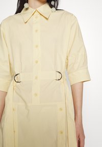 Ljust gult skjortklänning med en strukturerad krage, framknappar och bälte i midjan med silverdetaljer. Korta ärmar och slät tyg.