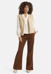 Beige faux-fur vest over a white blouse, paired with brown corduroy flared pants and cream heeled mules, featuring textured fabrics.