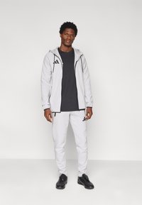 Γκρι φούτερ με φερμουάρ και μαύρες λεπτομέρειες, συνδυασμένο με μαύρο πουκάμισο και γκρι παντελόνια, με το λογότυπο της Adidas. Μαύρα παπούτσια ολοκληρώνουν το σύνολο.