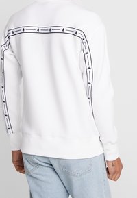 Persona che indossa un felpa bianca con strisce nere a motivo sulle spalle e lungo le maniche, abbinata a jeans di denim azzurro chiaro.