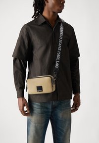 Bežová kožená crossbody taška so zipsom, doplnená štvorcovým logom z čierneho kovu a vzorovaným popruhom s bielym textom.