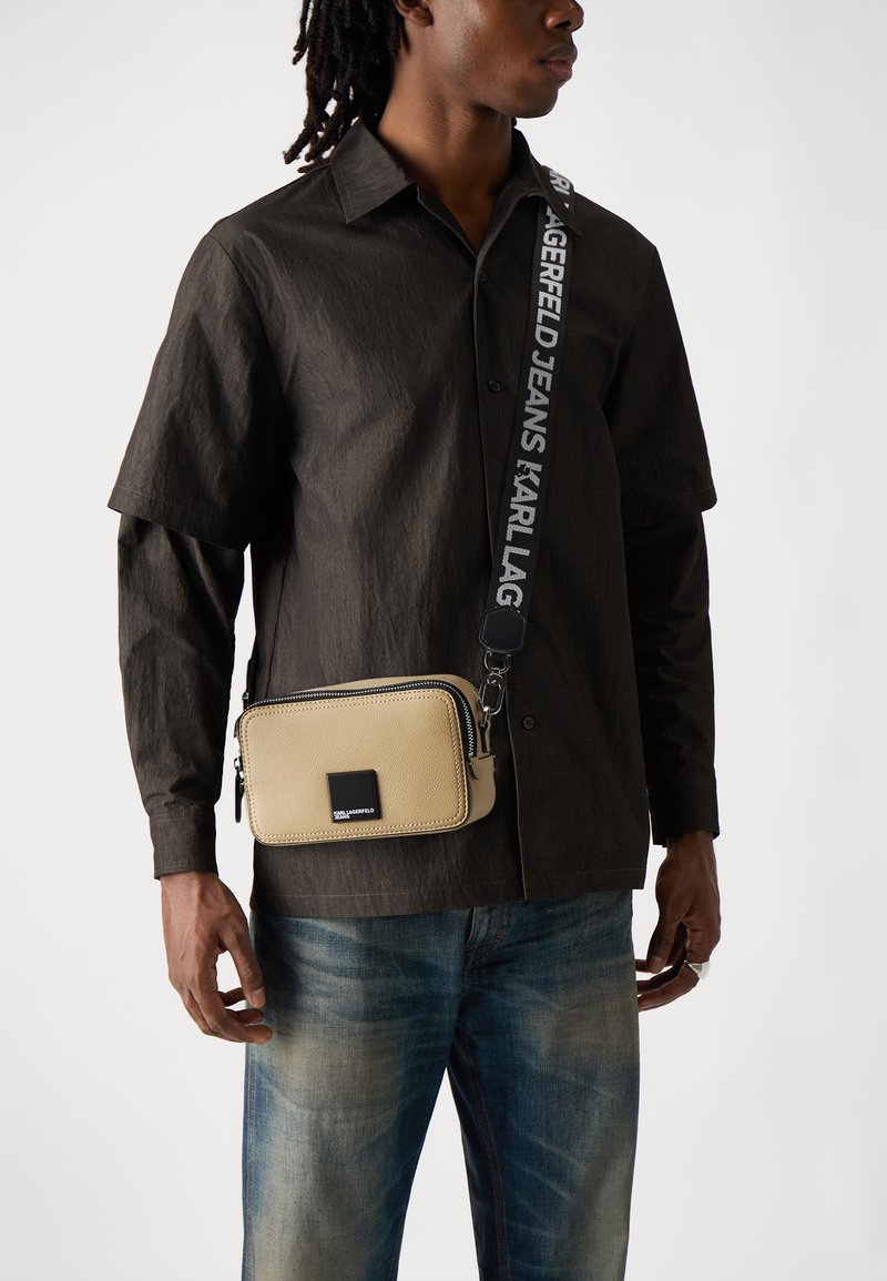 Bežová kožená crossbody taška so zipsom, doplnená štvorcovým logom z čierneho kovu a vzorovaným popruhom s bielym textom.