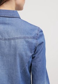 Chemise en denim avec un aperçu dorsal, à col, de couleur bleu clair et présentant une légère décoloration. Couture visible le long des coutures.