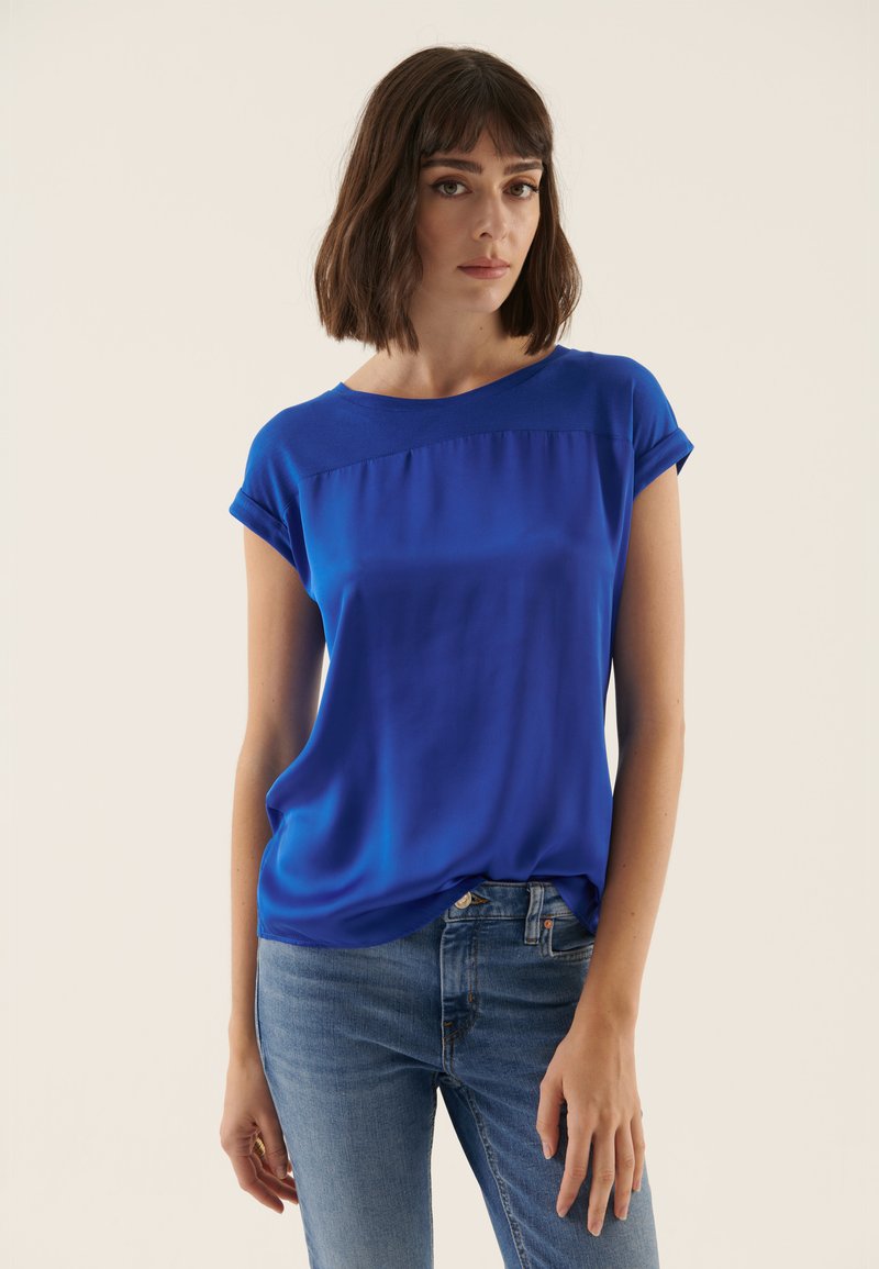 Blauwe blouse met korte mouwen, een gladde textuur en een ronde halslijn; heeft een lichte glans en plooitjes bij de buste. Gecombineerd met spijkerbroek.