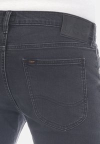 Jeans de denim negro con una textura suave, que presentan una etiqueta de cuero en la cintura, un pequeño logo naranja y detalles estándar en los bolsillos traseros.