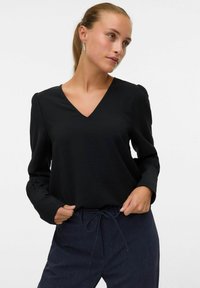Črn dolgo-sleeve top z V-izrezom, narejen iz teksturirane tkanine. Ima napihnjene ramena in ohlapen kroj. V kombinaciji s temno modrimi hlačami na vrvico.