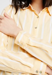 Camisa amarilla a rayas con líneas verticales blancas, que presenta un diseño de botones, cuello y puños con botones. Textura de tejido suave.