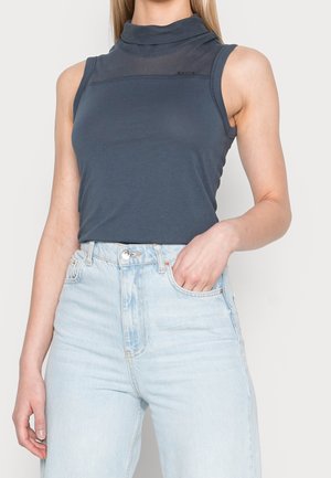 Vrouw draagt een mouwloze donkerblauwe coltrui top, ingestopt in een lichtblauwe jeans met hoge taille, één hand in de zak, tegen een witte achtergrond.