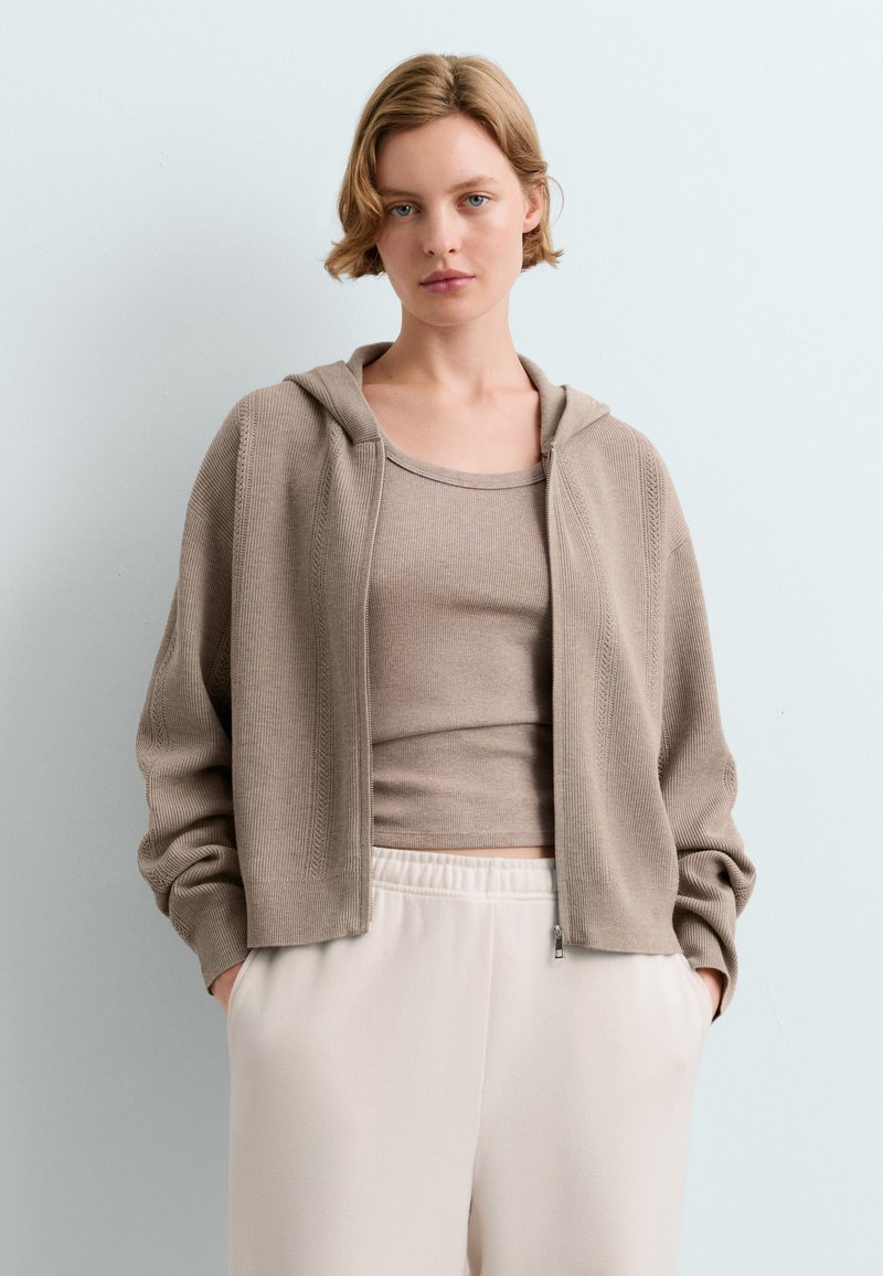 Jeune femme portant un cardigan à capuche beige zippé, un débardeur assorti et un pantalon blanc, les mains dans les poches, sur un fond clair uni.