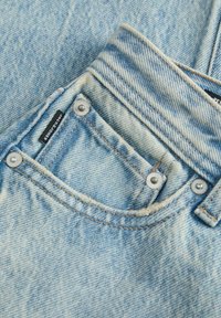 Jeans de mezclilla azul claro con un pequeño bolsillo frontal, detalles en botón a presión plateado y una etiqueta oscura cosida en la tela. Acabado texturizado.