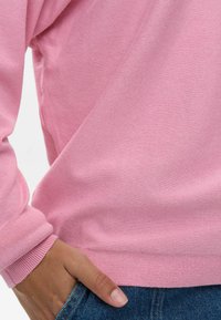 Suéter de punto rosa claro con un dobladillo y puños acanalados, mostrando una mano descansando en el bolsillo de unos vaqueros, destacando la textura suave y el ajuste casual.