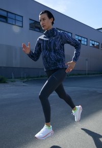 Kobieta w ciemnym stroju sportowym i białych butach do biegania biega na świeżym powietrzu w pobliżu budynków przemysłowych pod jasnym niebem.