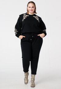 Zwarte sweater met bloemenborduurwerk op de mouwen, gecombineerd met zwarte joggers en enkellaarsjes met slangenprint. Zachte stof, ontspannen pasvorm.