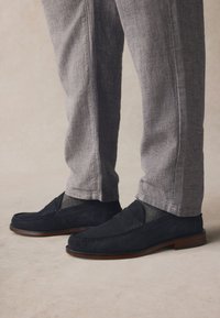 Marineblaue Wildleder-Loafer mit flachem Absatz, versehen mit einer gesteppten Verzierung über dem Spann. Kombiniert mit hellgrauen, strukturierten Hosen.