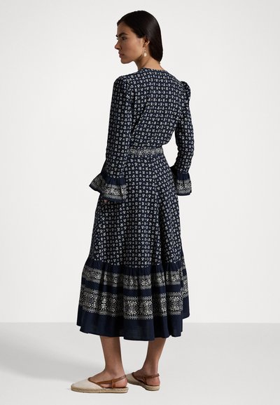 Laulu tummansininen mekko, jossa on valkoinen kukkakuosi, pitkät hihat, leveät hihansuut ja kerroksellinen hameosa. Kangas näyttää kevyeltä ja virtaavalta.