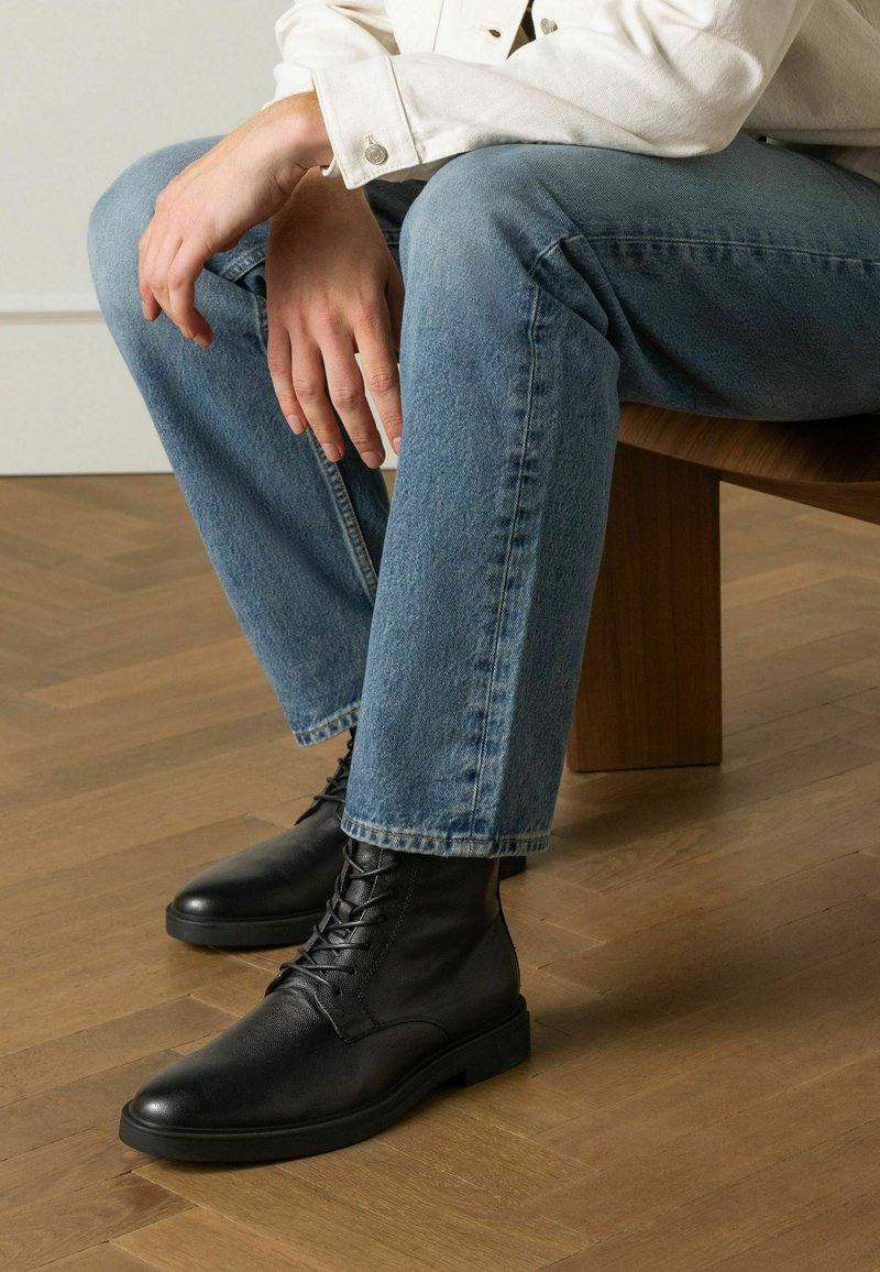Bottines en cuir noir avec un bout rond et un laçage à l'avant, associées à un jean en denim bleu clair, assises sur un banc en bois.