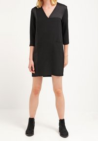 Robe noire avec un décolleté en V et des manches trois-quarts, présentant des sections de tissu mates et brillantes en contraste. Portée avec des bottines noires.
