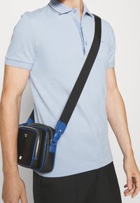 Světle modré polo tričko s krátkými rukávy a černým lemem, spárované s kompaktní černou a modrou křížovou taškou s dvojitými zipy.