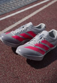 Harmaat juoksukengät, joissa on voimakkaat vaaleanpunaiset raidat, kevyestä verkkomateriaalista ja tyylikkäästä muotoilusta. Varustettu piikeillä, jotka tarjoavat pitoa radan pinnoilla.