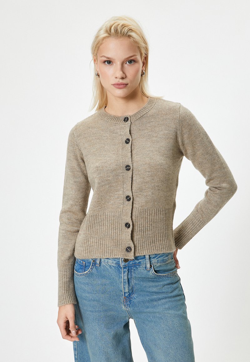 Cardigan lavorato a maglia beige con collo rotondo e otto bottoni scuri, con dettagli a costine ai polsi e all'orlo. Abbinato a jeans in denim blu.