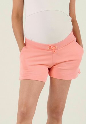 Schwangere Person, die ein weißes Oberteil und korallrosa Umstandsshorts mit Kordelzug trägt, die Hände in den Taschen hat.